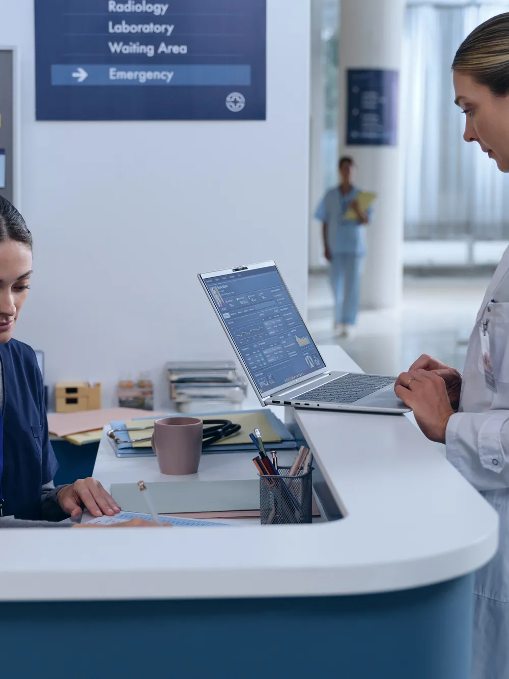 Clinicians at a hospital reception desk working on a laptop beside wayfinding signage